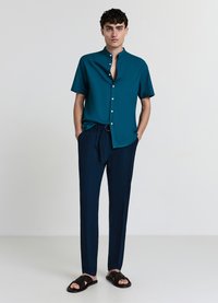 Chemise à manches courtes turquoise avec un col montant et des boutons blancs, assortie à un pantalon large bleu marine et des sandales noires à brides texturées.