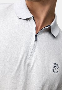 Polo grigio con colletto zip, caratterizzato da accenti blu navy e un logo ricamato. Tessuto morbido con una superficie liscia.