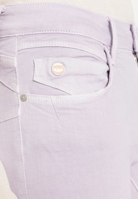 Jeans de ganga lilás claro com uma textura suave. Apresenta um bolso frontal com um botão de marca e costura em contraste.