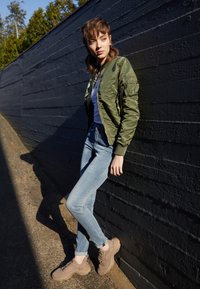 Giubbotto bomber verde oliva con tasche con zip, t-shirt bianca, jeans skinny azzurri chiari e sneaker color beige contro un muro scuro.