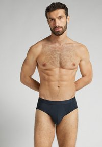 Svarta korta underkläder med en slät, stretchig yta och en åtsittande design. Bärs av en man med muskulös kropp och kort, mörkt hår.