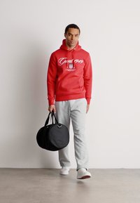 Homme en sweat à capuche rouge et pantalon de jogging gris marchant, tenant un sac de sport noir en forme de rond dans sa main droite sur un fond uni.