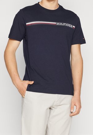 Mężczyzna w granatowej koszulce Tommy Hilfiger z czerwono-biało-niebieskim paskiem i białych spodniach, ręka w kieszeni, na jednolitym tle.