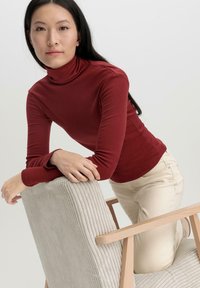 Burgunderfarbener Rollkragenpullover aus weichem Stoff, figurbetontes Design. Das Modell lehnt an einem strukturierten, hellbeigen, gerippten Stuhl mit Holzarmen.