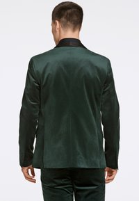 Blazer verde scuro, strutturato con una finitura liscia, presenta un risvolto nero, uno spacco singolo sul retro e polsini con bottone.