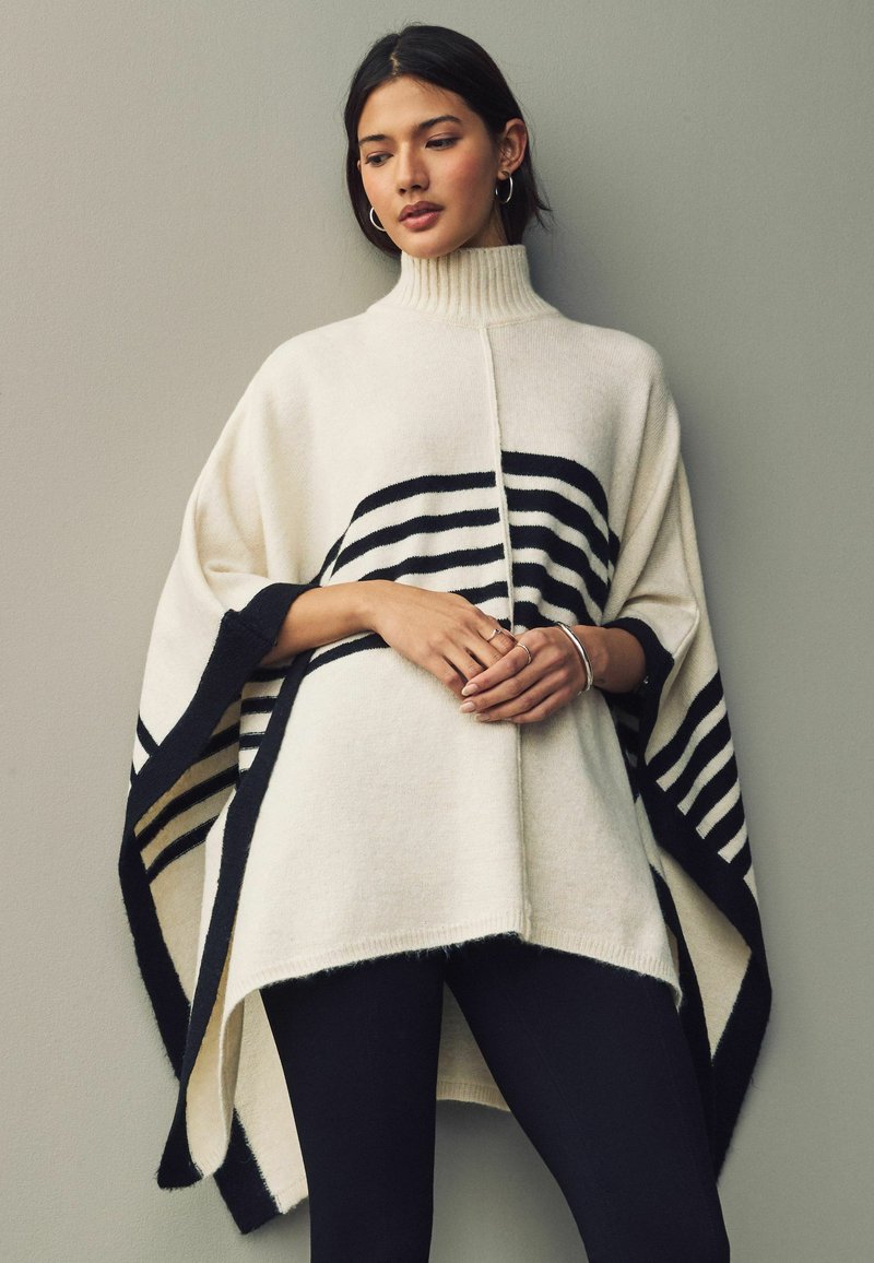 Cream-colored poncho featuring a high collar, black stripes, and a relaxed fit. Made of soft material with open sides and a contrasting black trim.