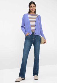 Helles lila geripptes Strickjacke mit braunen Knöpfen, getragen über einem gestreiften creme- und beigen Oberteil, kombiniert mit blauen Schlagjeans und cremefarbenen Mokassins.