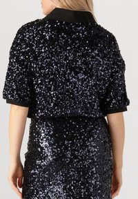 Vrouw in een zwart pailletten jurkje met korte mouwen en een kraag, van achteren zichtbaar tegen een effen achtergrond.