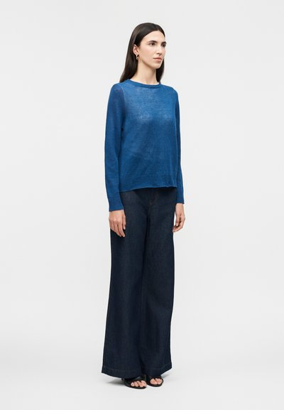 Femme aux longs cheveux bruns portant un pull bleu transparent, un jean large foncé et des sandales noires à bouts ouverts, debout devant un fond lumineux uni.