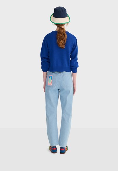 Mujer de pie de espaldas, vistiendo un suéter azul, jeans de mezclilla claros con un parche colorido, un sombrero de cubo bicolor y sandalias multicolores.