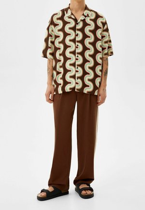 Brown short-sleeve shirt with wavy multicolor pattern, paired with loose brown pants and black slide sandals. Smooth, lightweight fabric.