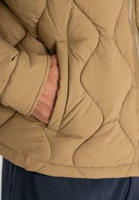 Beige takki, jossa on tyylikäs polyesterivuori, sivutaskut ja kontrastoiva ommel; varustettu vetoketjulla ja rentoa istuvuutta.