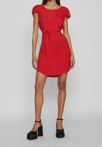 Vestido corto rojo con mangas cortas con volantes, cintura con cordón y dobladillo asimétrico. Llevado con tacones de plataforma negros. Textura de tela suave.