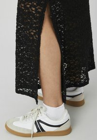 Robe en dentelle noire avec une fente haute, révélant une chaussette claire et une chaussure blanche avec des accents noirs et une semelle gomme, mettant en valeur les textures superposées.