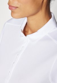 Polo shirt blanc avec une texture douce, doté d'un col classique, d'une patte de boutonnage et de coutures discrètes le long des épaules.