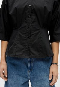 Gros plan sur une personne portant une chemise noire boutonnée à manches 3/4, un jean en denim bleu, et un bracelet argenté au poignet droit.