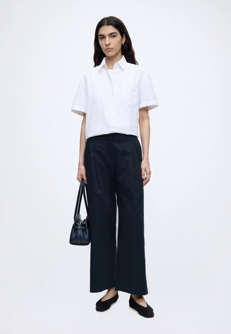 Femme aux longs cheveux foncés portant une chemise blanche à manches courtes boutonnée, un pantalon noir à jambes larges, des ballerines noires et tenant un sac à main noir.