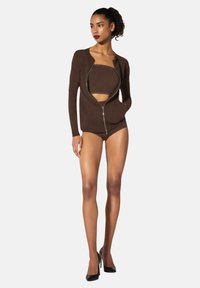 Brun zip-up jacka över en matchande bandeau-top och shorts, allt i ett mjukt tyg. Modellen har på sig svarta högklackade skor, vilket framhäver en elegant silhuett.