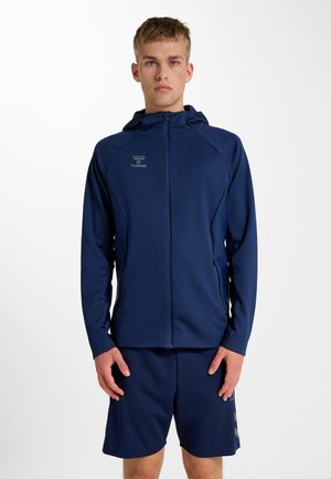 Young man wearing a navy blue hooded zip-up jacket and matching shorts, standing against a plain white background.