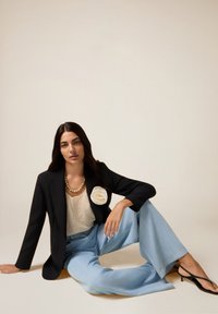 Frau, die auf dem Boden sitzt, trägt ein schwarzes Blazer mit einer weißen Blumenbrosche, ein cremefarbenes Strickoberteil, hellblaue weitem Hosen und schwarze hohe Sandalen.