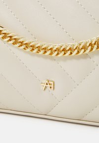 Beige quiltet taske med en guld kædestrop og prægede logo. Glat tekstur med skrå syninger og gulddetaljer.