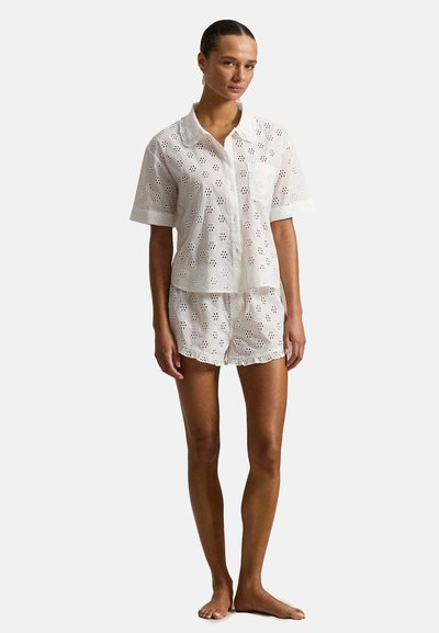 Vrouw staat op blote voeten, draagt een wit shirt met korte mouwen en knoopjes, en bijpassende shorts met bloemenpatronen in opengewerkt geborduurd kant.