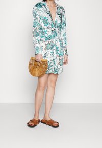 Biała sukienka w kwiatowy wzór z wiązaniem z przodu, w zestawie z okrągłą torbą ze słomy bambusowej i brązowymi sandałami z frędzlami. Gładki materiał, luźny krój.