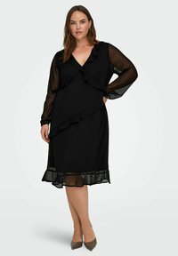Czarna sukienka z falbaniastymi akcentami, długimi przezroczystymi rękawami i rozkloszowanym hemem. Wykonana z miękkiego materiału, z głębokim dekoltem w kształcie V.
