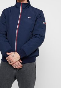 Mörkblå bomberjacka med hög krage och röd och vit dragkedja. Har ribbade manschetter och en liten logotyp på bröstet.