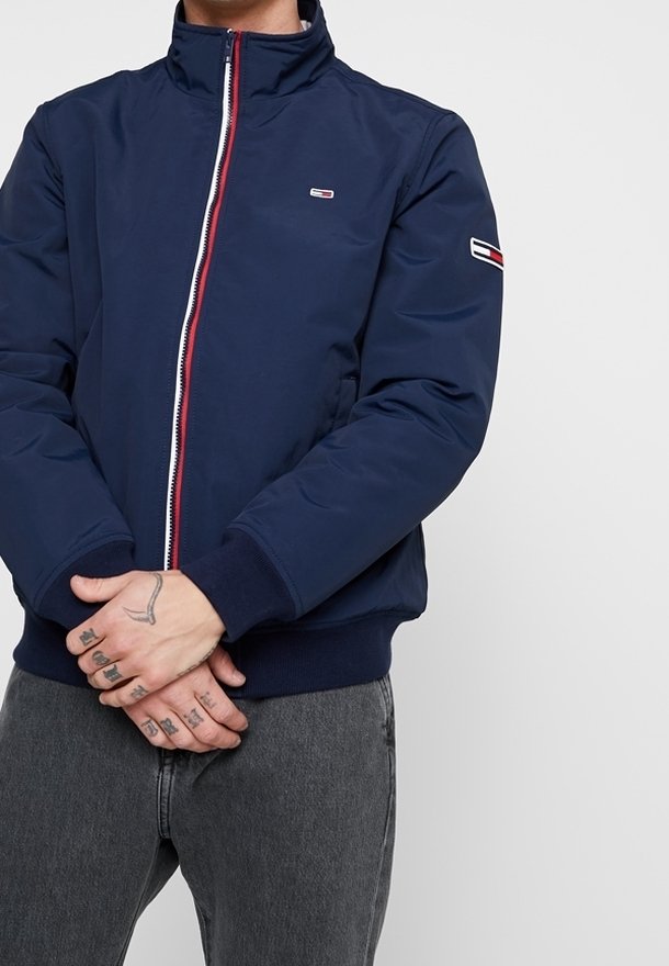 Mörkblå bomberjacka med hög krage och röd och vit dragkedja. Har ribbade manschetter och en liten logotyp på bröstet.