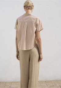 Camicetta a maniche corte di un beige chiaro con strisce verticali blu e lavanda, abbinata a pantaloni beige a gamba larga, vista da dietro.