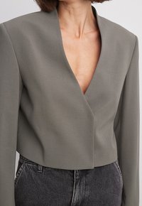 Giacca grigia corta con scollatura profonda a V, spalle strutturate e tessuto liscio, abbinata a jeans neri a vita alta.