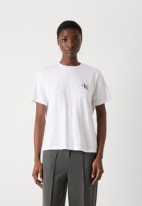 White cotton t-shirt featuring a black logo on the chest, round neckline, and short sleeves, paired with gray tailored pants.