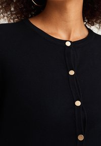 Svart kofta med rund halsringning, prydd med guldtonade knappar fram och en slät, strukturerad tyg.