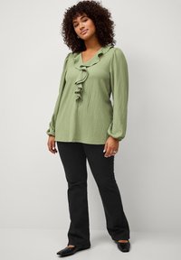 Blouse vert clair à manches longues avec un col volanté, en tissu texturé, associée à un pantalon noir évasé et des chaussures plates.