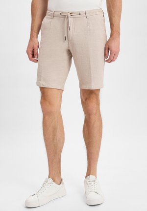 Beige Leinen-Shorts mit einem Kordelzugbund, seitlichen Taschen und umgeschlagenen Säumen. Kombiniert mit weißen Sneakers vor einem hellen Hintergrund.