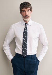 Camicia bianca con colletto a punta, abbinata a una cravatta blu scuro con piccoli pois chiari e pantaloni a righe sottili scuri.