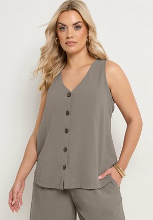 Blonde Frau trägt ärmelloses graues Oberteil mit Knöpfen und dazu passende Hose, dazu goldene Creolen und Armbänder, posiert vor einfarbigem Hintergrund.