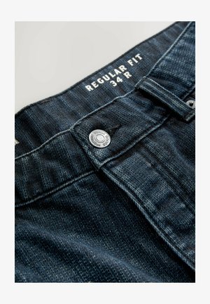 Reguliaraus kirpimo džinsai tamsiai mėlynos spalvos, su sidabriniu mygtuku ir kontrastinga siūle. Audinys turi švelnią tekstūrą ir lengvą nusidėvėjimą.