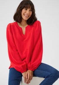 Blouse rouge avec un décolleté en V et des manches volumineuses, dotée d'un motif rayé texturé. Associée à un jean en denim bleu.