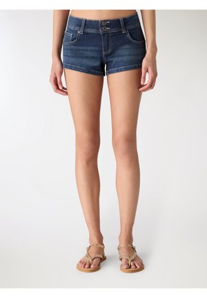 Kobieta w ciemnoniebieskich dżinsowych szortach z podwójnym zapięciem na guziki i beżowych płaskich sandałach, stojąca na jednolitym białym tle.