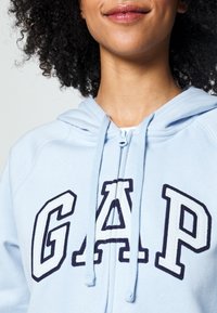Light blue hoodie featuring a zip-up front, adjustable drawstring, and bold navy "GAP" lettering in a stitched design.