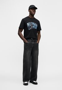 Μαύρο βαμβακερό t-shirt με γαλάζια γραφική γραφή, συνδυασμένο με φαρδιά παντελόνια από ξεθωριασμένο μαύρο τζιν με διακριτικές λεπτομέρειες ραφής. Μαύρο καπέλο και παπούτσια.