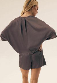 Kobieta z blond włosami, ubrana w luźną, krótko rękawową bluzkę w kolorze taupe i pasujące szorty, stoi z jedną ręką na biodrze, odwrócona tyłem.