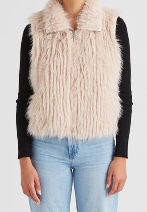 Woman wearing a light beige fluffy sleeveless zip-up vest over a black long-sleeve top and light blue jeans.