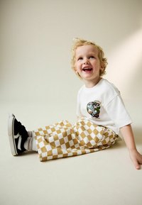 Camiseta blanca de manga corta con estampado gráfico, combinada con pantalones de pierna ancha a cuadros amarillos y blancos. Zapatos negros con suelas blancas.