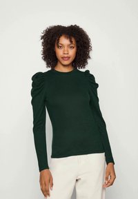 Top de manga longa, verde escuro, com ombros volumosos, decote redondo e uma textura suave, combinado com calças de cor clara.