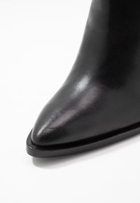 Černá kožená kotníková bota s špičatou špičkou, hladkou texturou a minimalistickým designem. Obsahuje nízký profil podrážky pro větší stabilitu.