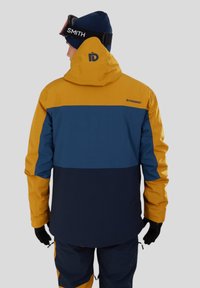 Veste d'hiver colorée avec capuche, présentant des panneaux jaunes, marine et bleu foncé. Fabriquée en matériau texturé et imperméable. Manches ajustables.