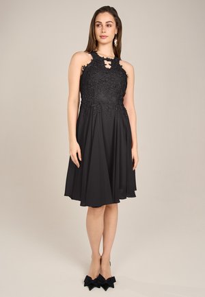 Schwarzes Cocktailkleid mit einem Spitzenoberteil und floralem Stickerei, ärmelloses Design und einem schwingenden Rock. Kombiniert mit schwarzen, mit Schleifen verzierten Absätzen.